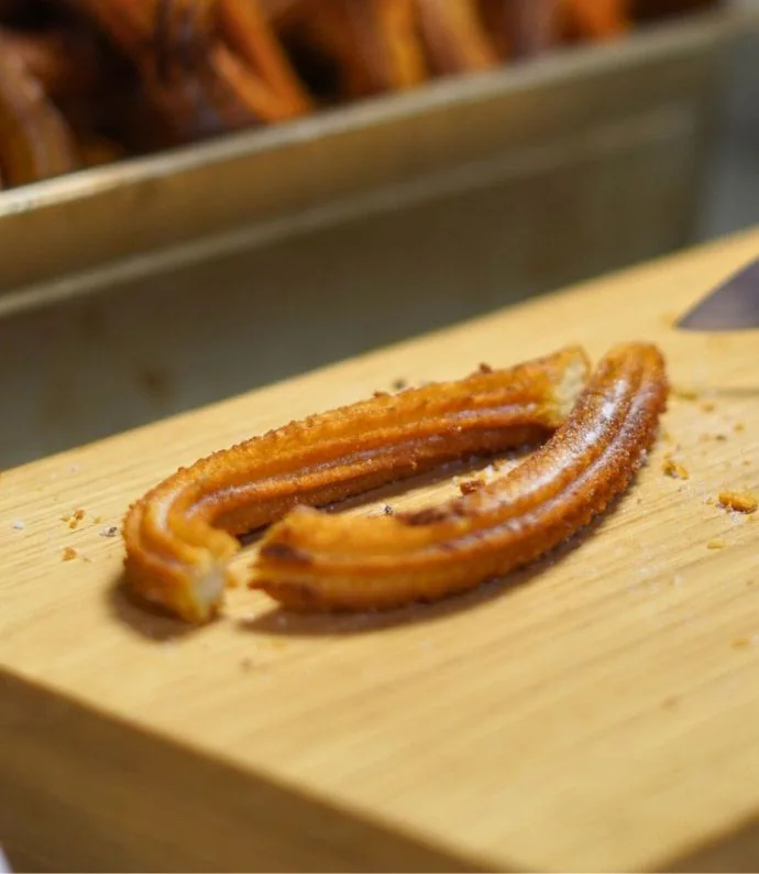 A freshly fried churro resting on a wooden board beside a tray of others