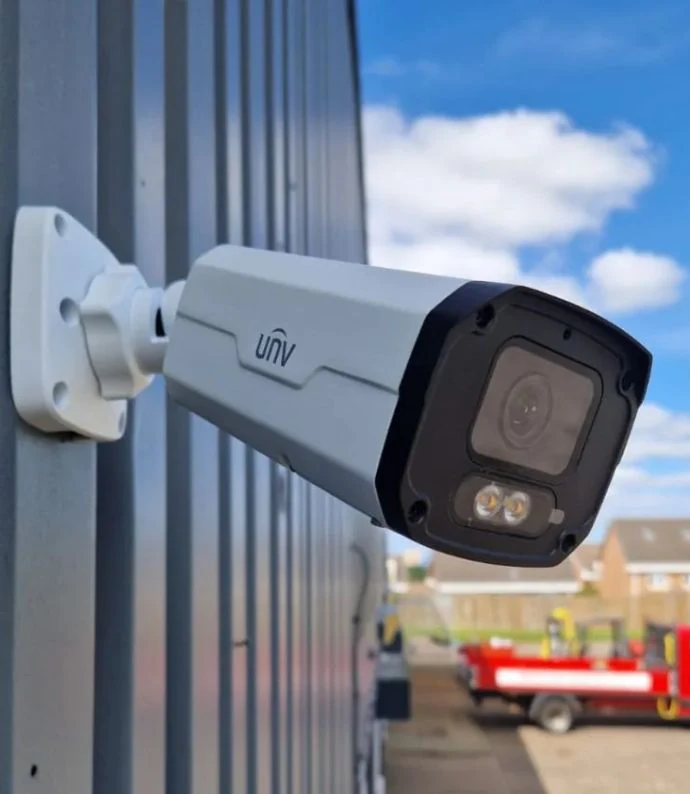 A wall mounted CCTV camera on the side of a steel clad building, overlooking a yard