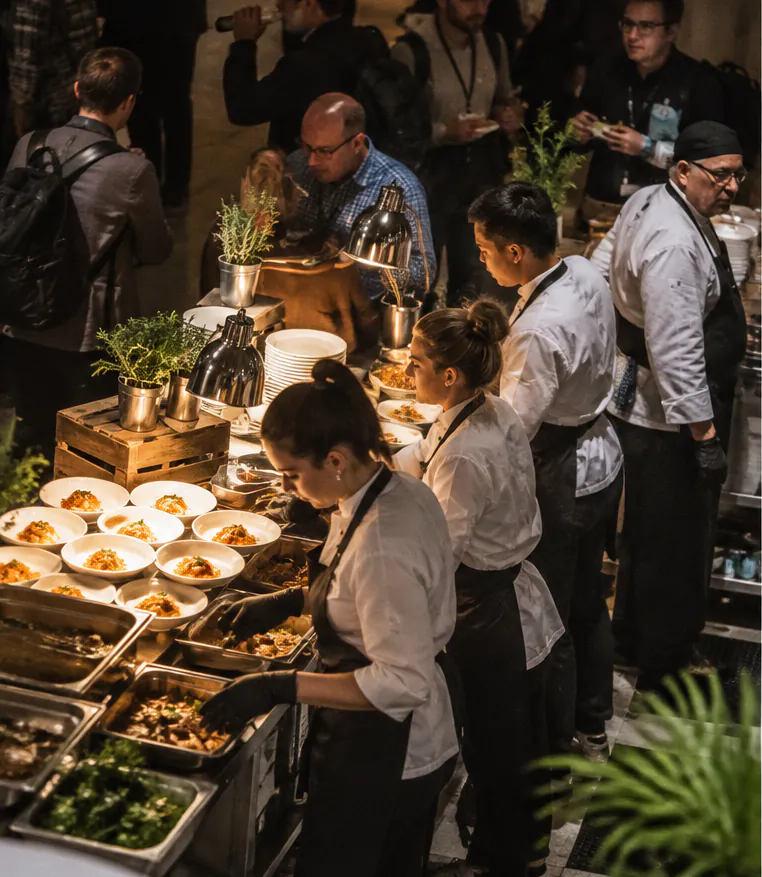 Catering staff in white shirts and aprons serving dishes at a busy event buffet